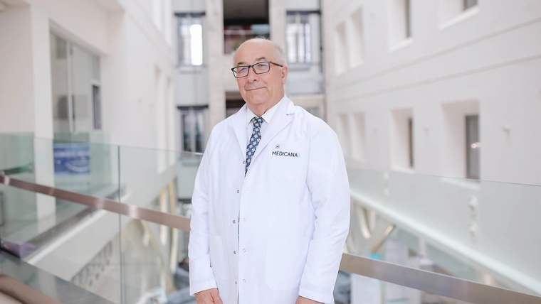 Medicana yazılı beyaz önlük giyen gözlüklü bir kişi, cam korkuluklu aydınlık bir koridorda duruyor.