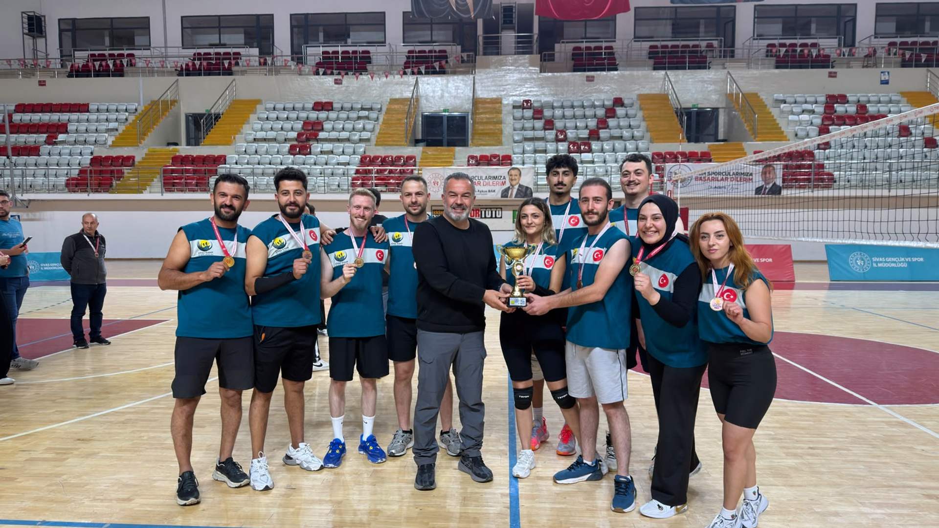 Voleybol sahasında madalya ve kupayla poz veren sporcular, ortalarında bir kişiyle birlikte duruyor.