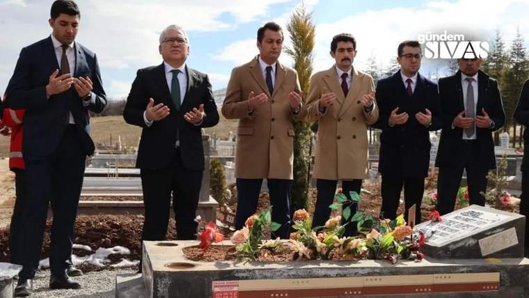 Mezarlıkta altı erkek, çiçeklerle süslenmiş bir mezarın önünde ellerini açmış duruyor.