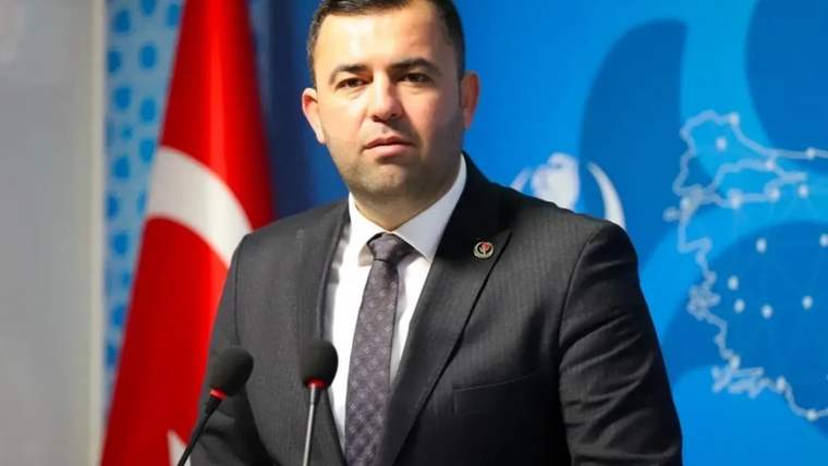 Takım elbiseli bir kişi, Türk bayrağı ve harita desenli pano önünde iki mikrofonla konuşuyor.