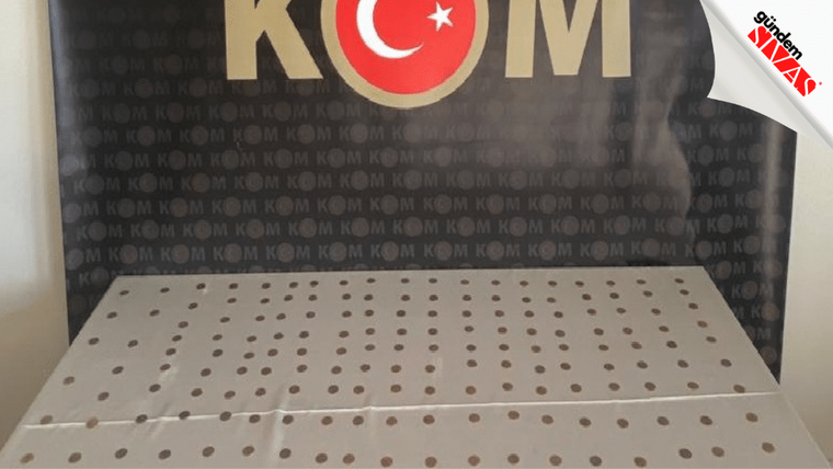 Üzerinde çok sayıda delik bulunan metal bir tabla, arkasında KOM yazılı bir pano önünde duruyor.