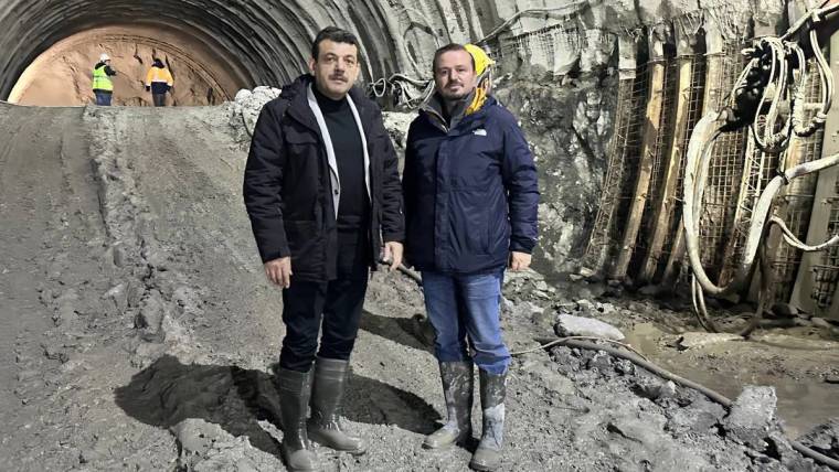 Tünel içinde iki kişi, kazı alanındaki kaya yüzeyi ve destek ekipmanlarının önünde duruyor.