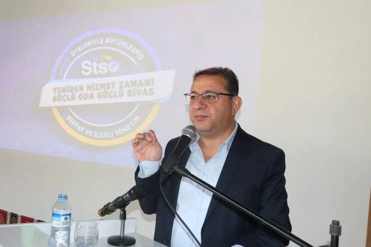 Mikrofon başında konuşan gözlüklü bir adam, elini kaldırıyor; arkasındaki yansıtılan görselde Sivas yazıyor.