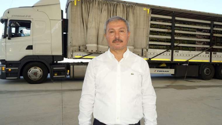 Beyaz gömlekli bıyıklı bir kişi, kasalarla yüklü bir kamyonun önünde kapalı alanda duruyor.