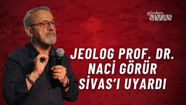 Mikrofon tutan gözlüklü bir kişi, kırmızı harita fonu ve büyük beyaz yazılar önünde duruyor.