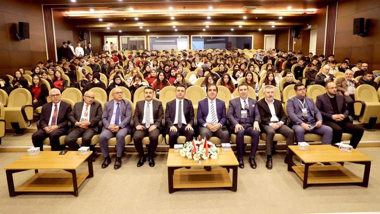 Amfi salonunda önde takım elbiseli kişiler, arkalarında ise sıralara oturmuş kalabalık bir grup görülüyor.