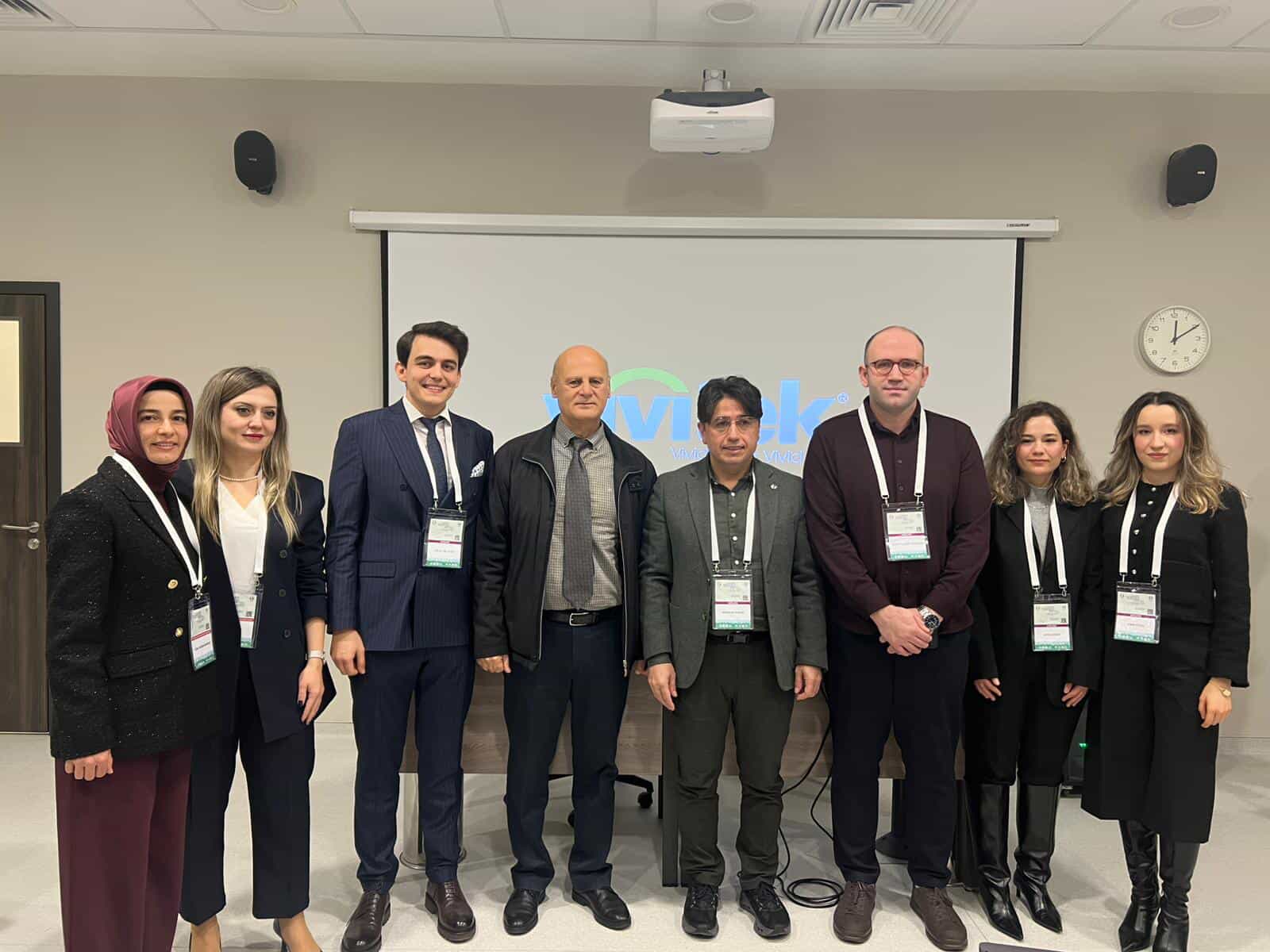Toplantı salonunda, projektör perdesinin önünde duran sekiz kişi yan yana poz veriyor.