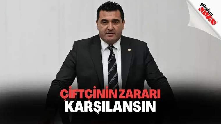 Kravatlı takım elbiseli bir kişi kürsüde konuşurken, önünde “ÇİFTÇİNİN ZARARI KARŞILANSIN” yazısı görülüyor.