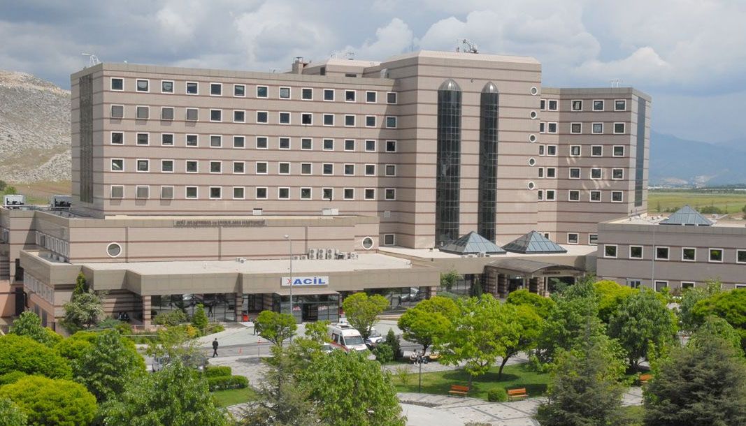 Çok katlı hastane binasının girişinde “ACİL” tabelası, önünde ağaçlı bir alan ve araçlar görülüyor.