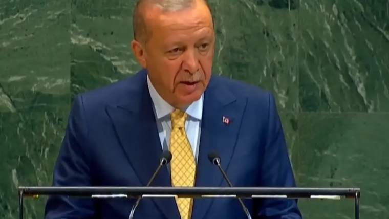 Mavi takım elbise giymiş bir kişi, podiumda mikrofonun önünde konuşuyor.