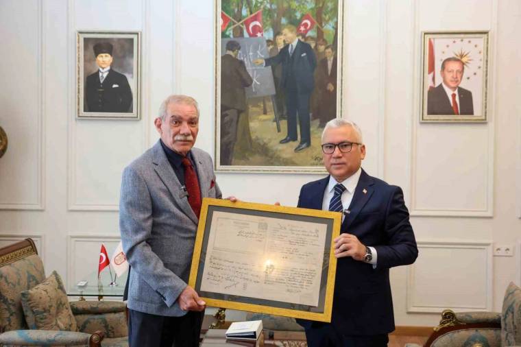 İki erkek, duvarda portreler ve yağlı boya tablo bulunan odada çerçeveli bir belge tutuyor.