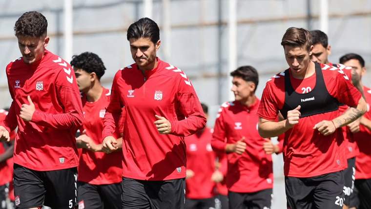 Kırmızı formalar giyen futbolcular, çim sahada toplu halde koşu antrenmanı yapıyor.