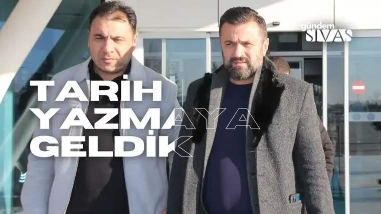 İki kişi, bir binanın önünde duruyor; biri siyah ceket, diğeri gri mont giymiş.