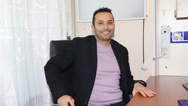 Bir ofiste oturan, gülümseyen bir erkek doktor, masanın arkasında duruyor.