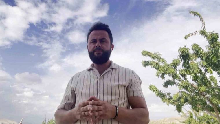 Açık havada duran sakallı bir kişi, çizgili gömleğiyle ellerini birleştirerek kameraya bakıyor.