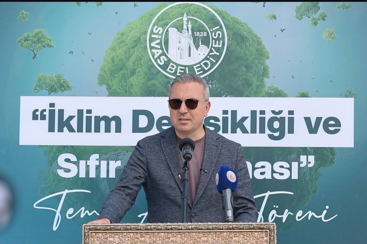 Sivas Belediyesi logolu pankart önünde, kürsüde konuşan bir kişi ve iki mikrofon görülüyor.