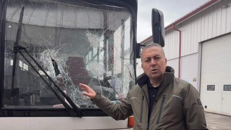 Bir kişi, camı kırılmış bir otobüsün önünde duruyor ve camı gösteriyor.