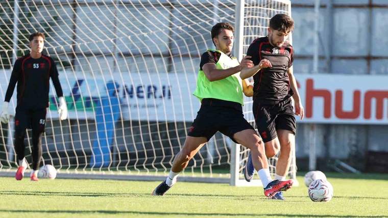 Futbol antrenmanında iki oyuncu top için mücadele ederken, kaleci ve kale ağı da arka planda görülüyor.