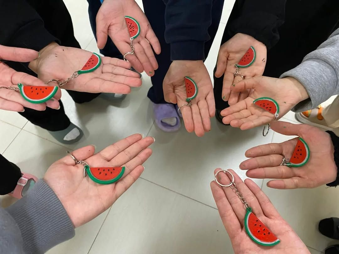 Bir grup kişinin avuçlarında kırmızı ve yeşil karpuz figürlü anahtarlıklar görülüyor.