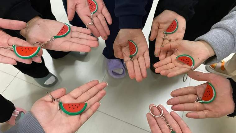 Bir grup kişinin avuçlarında kırmızı ve yeşil karpuz figürlü anahtarlıklar görülüyor.