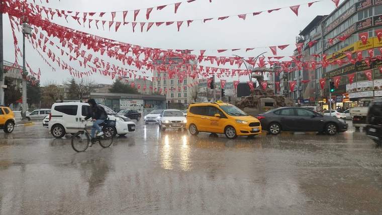Yağmur altında su birikintileriyle kaplanan kavşakta sarı taksi, otomobiller ve bisikletli ilerliyor.