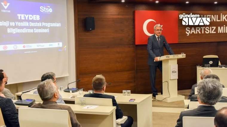 Kürsüde konuşan bir kişi, arkasındaki Türk bayrağı ve sunum ekranı önünde izleyicilere hitap ediyor.