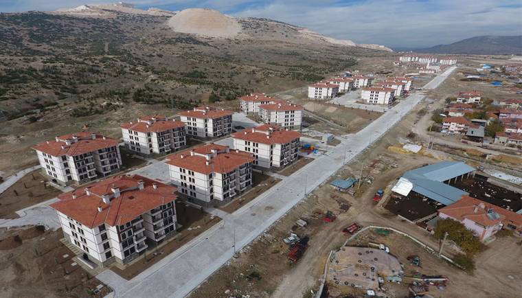 Dağlık arazinin eteklerinde, kırmızı çatılı çok katlı konut blokları ve çevre yolları görülüyor.