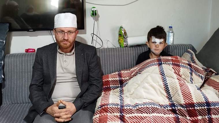 Bir adam, üzerinde beyaz bir başlık ve ceketle oturuyor; yanında bir çocuk, battaniyeye sarılı ve başında bandaj var.