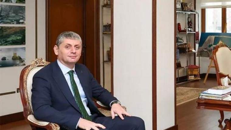 Koyu takım elbiseli bir erkek, çalışma odasında süslü bir koltukta oturuyor; arkada masa ve raflar bulunuyor.
