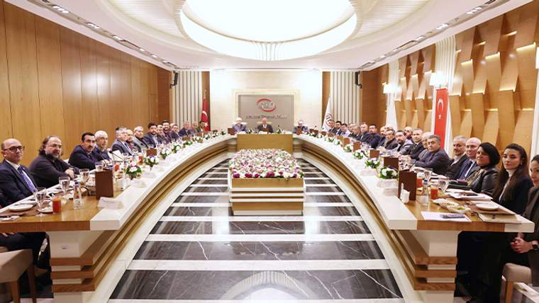 Geniş toplantı salonunda, U biçimli masada oturan kalabalık bir grup ve ortada çiçek düzenlemesi görülüyor.