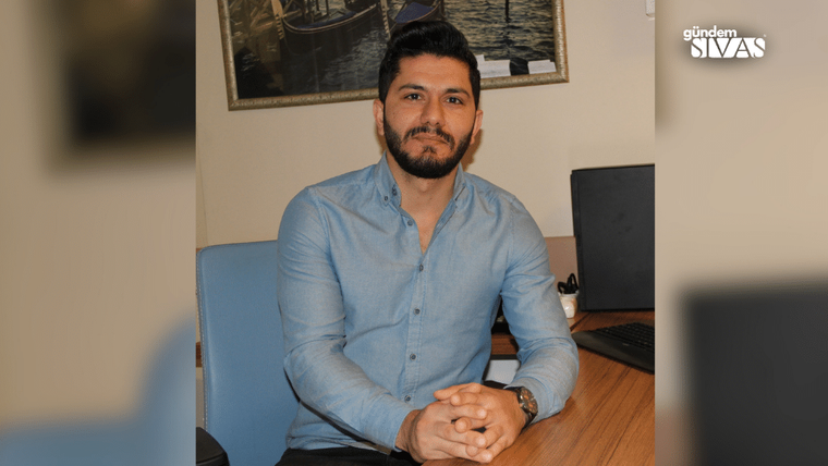 Masa başında oturan sakallı bir kişi, mavi gömlekle ellerini birleştirerek kameraya bakıyor.