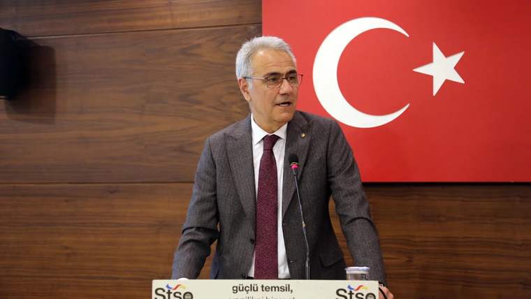 Kürsüde konuşan bir kişi, arkasında Türk bayrağı ve bir tabela bulunuyor.