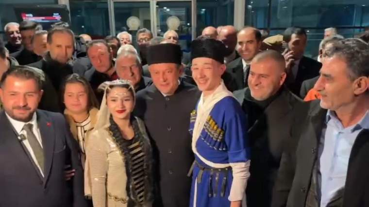 Kalabalık bir grup, cam giriş önünde poz verirken ortada mavi geleneksel kıyafetli bir kişi görülüyor.