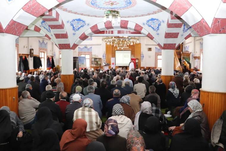Bir cami içinde, kalabalık bir topluluk bir sunumu dinliyor. Katılımcılar çeşitli kıyafetler giymiş.