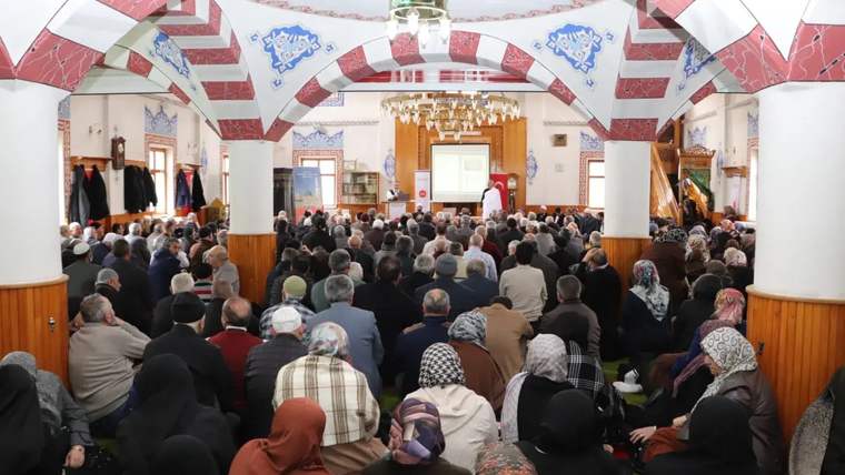 Bir cami içinde, kalabalık bir topluluk bir sunumu dinliyor. Katılımcılar çeşitli kıyafetler giymiş.