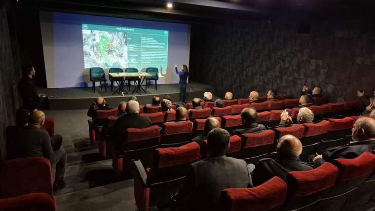 Bir sunum salonunda, bir kişi projeksiyon ekranında harita gösteriyor, izleyiciler dikkatle dinliyor.