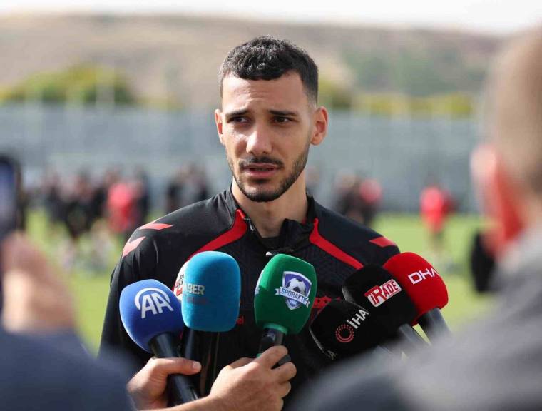 Siyah-kırmızı spor forması giyen bir kişi, açık alanda çok sayıda mikrofonun önünde konuşuyor.