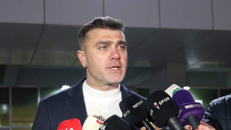 Mikrofonlarla çevrili bir kişi, bina girişinde basın mensuplarına açıklama yapıyor.