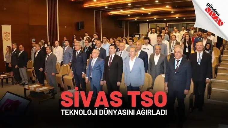 Konferans salonunda takım elbiseli ve günlük giyimli çok sayıda kişi sıralar arasında ayakta duruyor.