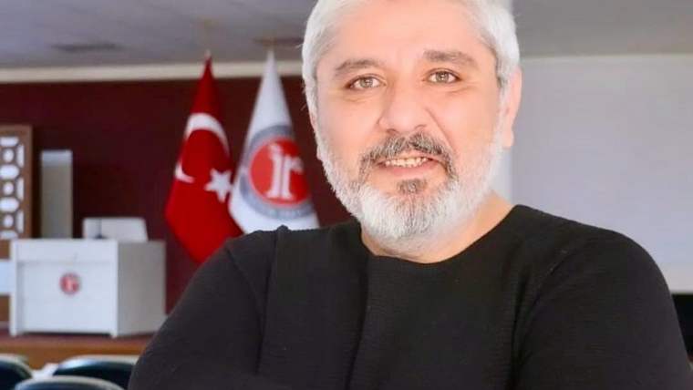 Beyaz saçlı ve sakallı bir erkek, arkasındaki Türk bayrağı ve amblemli beyaz bayrak önünde duruyor.
