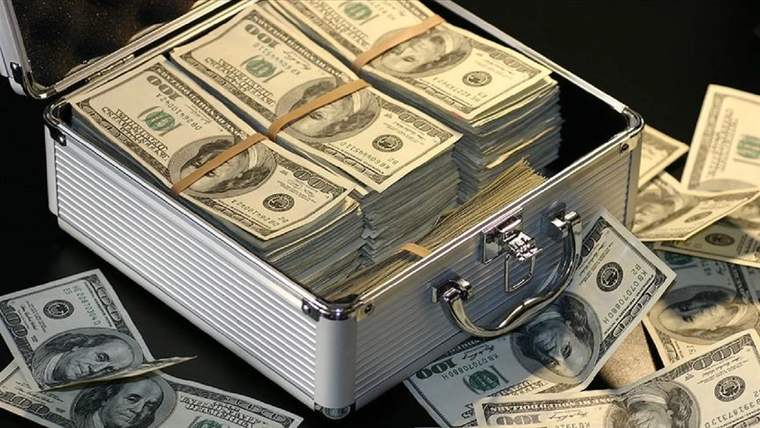Açık gümüş renkli bir çanta, deste halinde ABD doları banknotlarıyla dolu; çevresine de banknotlar saçılmış.