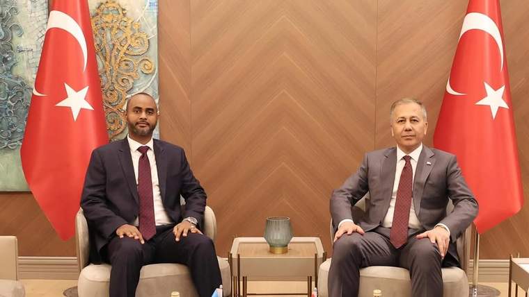 İki takım elbiseli kişi, Türk ve Somali bayraklarının bulunduğu salonda karşılıklı koltuklarda oturuyor.