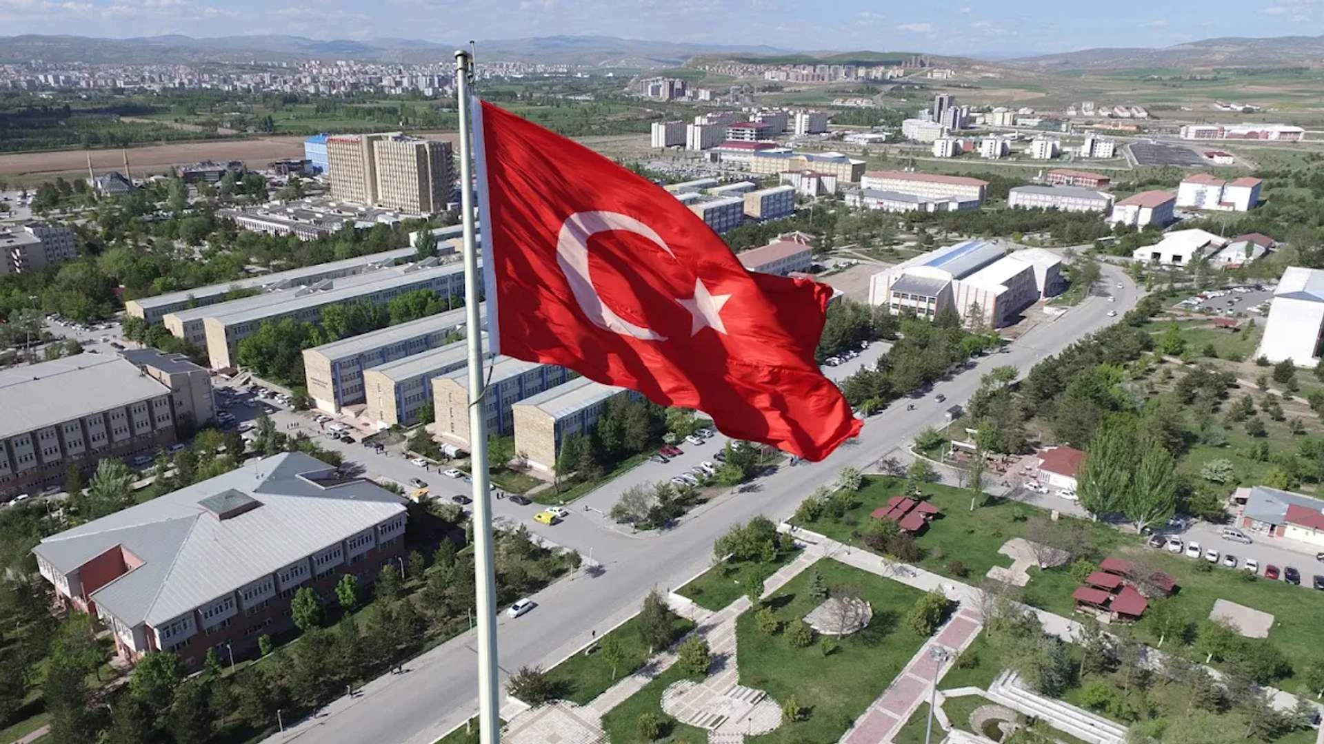 Ay yıldızlı Türk bayrağı, geniş bir yerleşkedeki direğinde dalgalanıyor; çevrede binalar, yollar ve ağaçlar görülüyor.