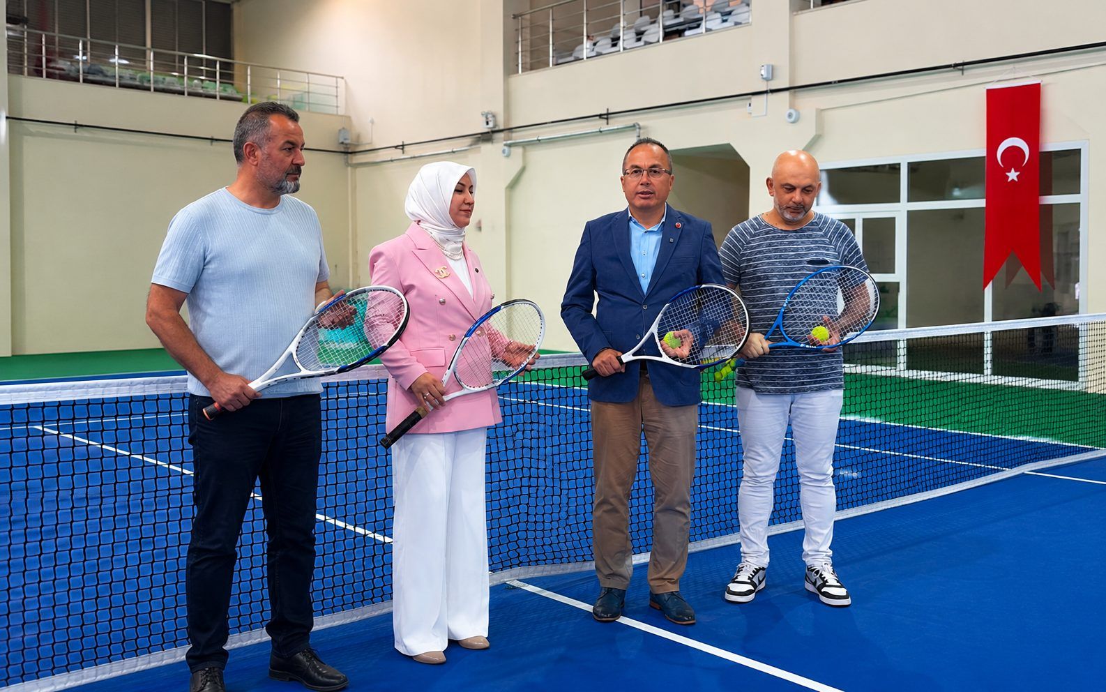 Kapalı tenis kortunda dört kişi raketleriyle file önünde duruyor; duvarda Türk bayrağı asılı.