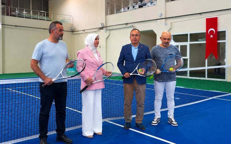 Kapalı tenis kortunda dört kişi raketleriyle file önünde duruyor; duvarda Türk bayrağı asılı.