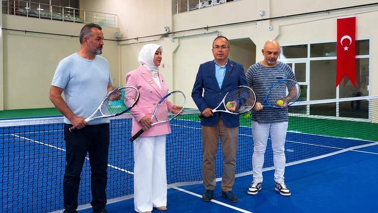 Kapalı tenis kortunda dört kişi raketleriyle file önünde duruyor; duvarda Türk bayrağı asılı.