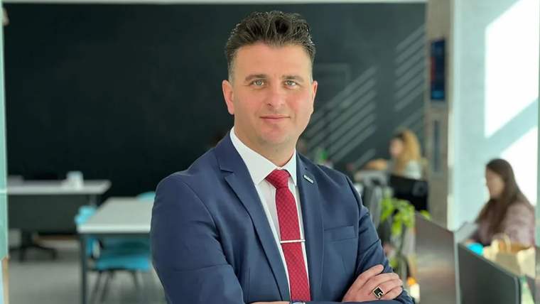 Kırmızı kravat takan, mavi takım elbise giymiş bir kişi, kollarını kavuşturmuş şekilde gülümsüyor.