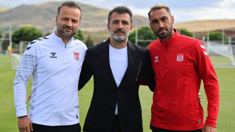 Futbol sahasında üç erkek yan yana duruyor; ortadaki siyah ceketli, diğer ikisi beyaz ve kırmızı spor kıyafetli.