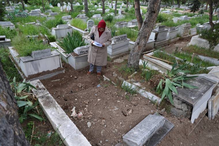 Bir kadın, mezarlıkta bir mezarın başında not alıyor. Etrafında mezarlar ve yeşil bitkiler var.