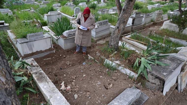 Bir kadın, mezarlıkta bir mezarın başında not alıyor. Etrafında mezarlar ve yeşil bitkiler var.